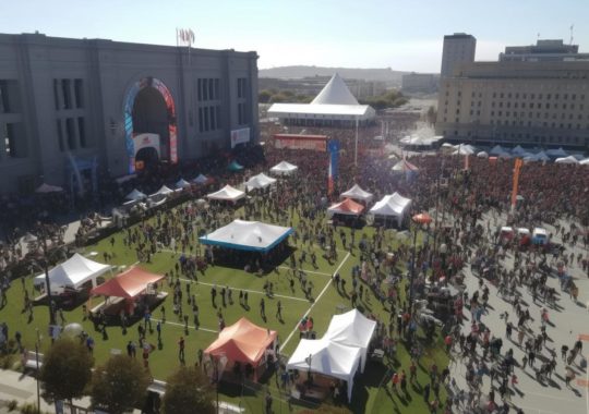 Una vista aérea de un festival bullicioso, con grandes pantallas y mesas, lleno de gente, evocando el estilo del Renacimiento de San Francisco, con tonos de azul claro, rojo, naranja oscuro y gótico del medio oeste.