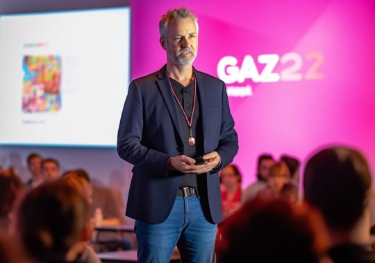 "Un hombre se encuentra frente a una audiencia en una conferencia animada, ilustrado en el estilo colorido de Lisa Frank y la precisión meticulosa de Carsten Meyerdierks, con una energía juvenil y tonos oscuros de plata y rosa, evocando la obra de Richard Gerstl."