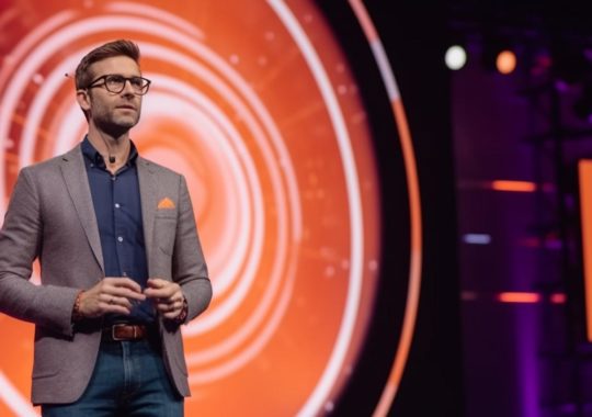 "Un hombre hablando frente a un fondo naranja brillante, con líneas radiantes y espirales en tonos grises oscuros y morados, evocando una espectacular muestra de épocas."