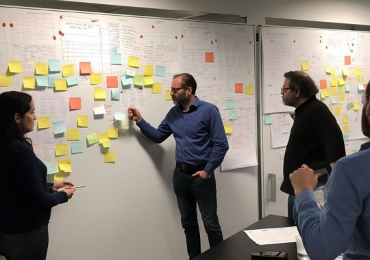 Un grupo de personas reunidas alrededor de una mesa, pegando notas en una pared, en un estilo de diseño equilibrado y líneas angulares, evocando una sensación de nueva objetividad y gorpcore.