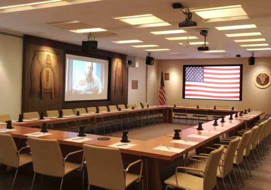 "Una sala de conferencias con una bandera americana en el fondo, presentada en tonos marrones claros y beige, con una disposición simétrica pero asimétrica, evocando técnicas tradicionales y un estilo manapunk en una construcción de hormigón armado."