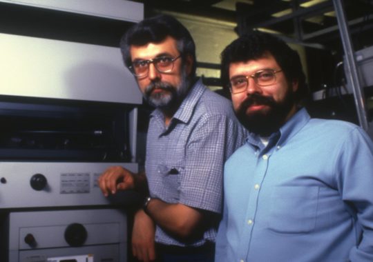 "Imagen de dos hombres junto a una máquina, en un estilo que entrelaza fotografía y materiales ópticos, evocando el arte de Jan Berenstain, Don Maitz y George Lucas."